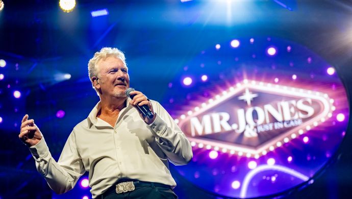 Mr Jones staat met een microfoon op een podium. Achter hem hangt een grote lamp met Mr. Jones erop.