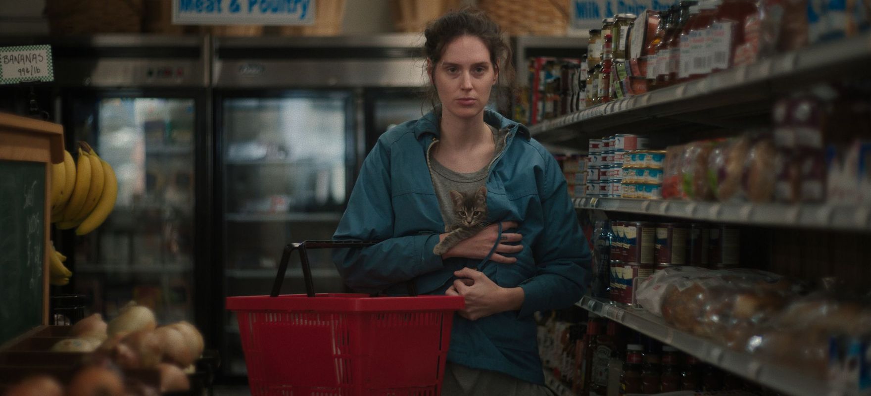 Vrouw in blauwe jas met daarin een kitten loopt met een rode winkelmand door een laan van een supermarkt. 