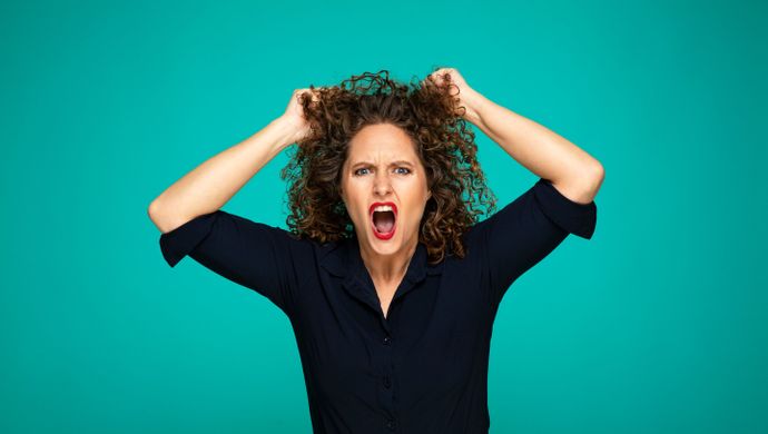 Een vrouw kijkt boos de camera in en heeft haar handen in haar haren. Ze heeft een blauwe blouse aan en rode lippenstift op. Ze staat voor een groene achtergrond