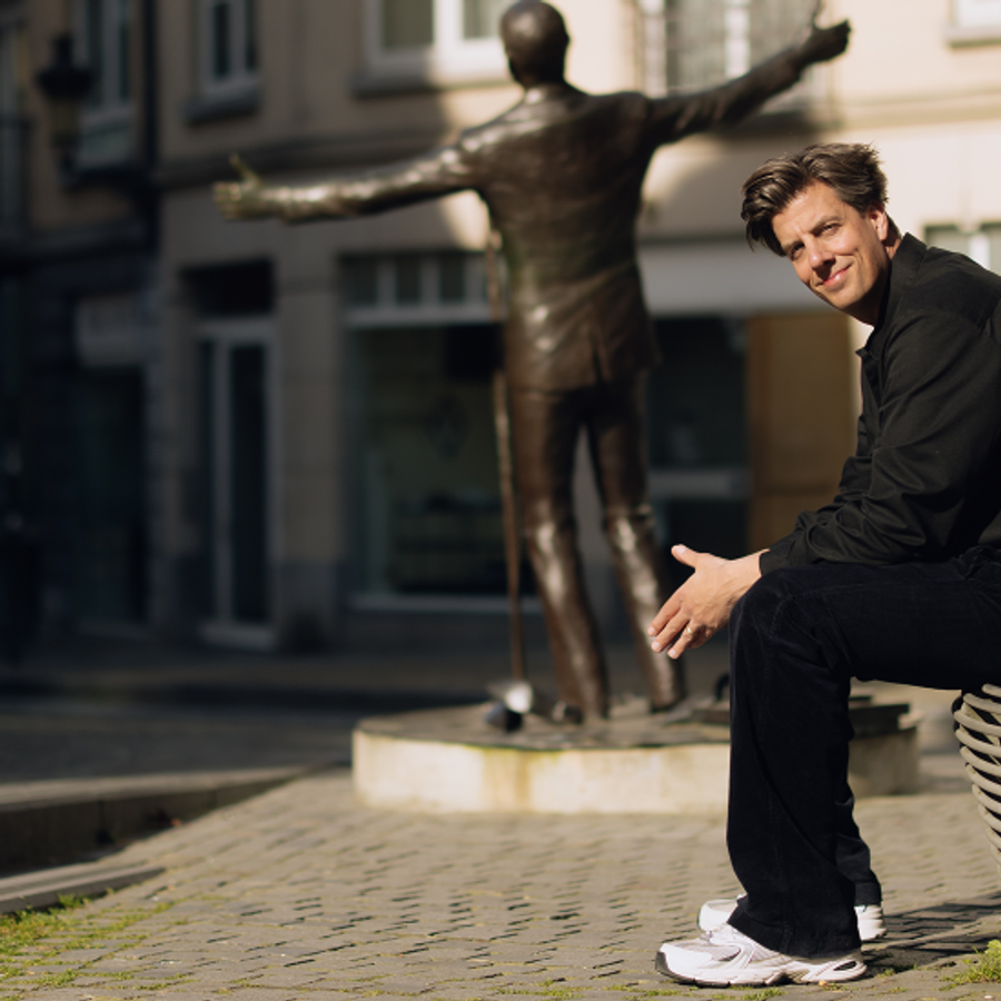 Rob kemps zit voorovergebogen op een bankje met zijn handen bij elkaar. De omgeving is een pleintje met een standbeeld in het midden. Hij kijkt glimlachend de camera in terwijl hij opzij kijkt.