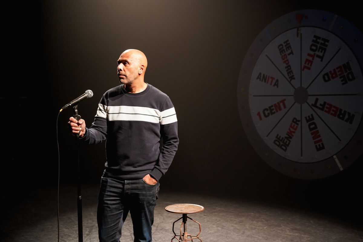 Man staand op een vrijwel donker podium voor een microfoon. 