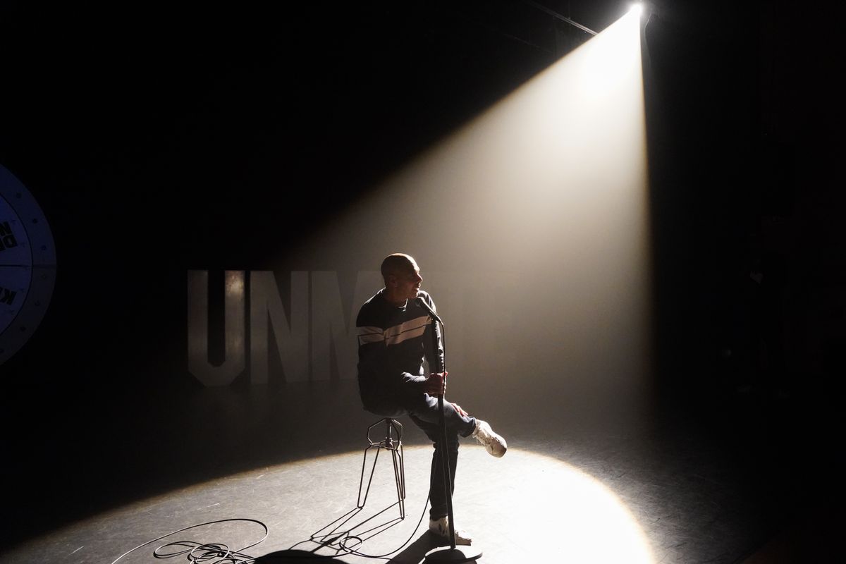 Man zittend op een stoel op een geheel donker podium met één spotlight op hem gericht. 