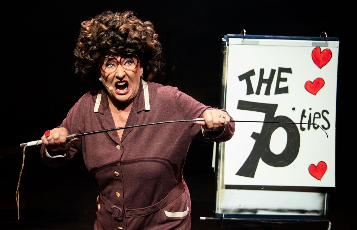 Karin bloemen heeft een zwarte pruik op en een pyjama aan. Ze houdt boos een stok in haar handen en naast haar staat een bord met: The 70'ties erop.