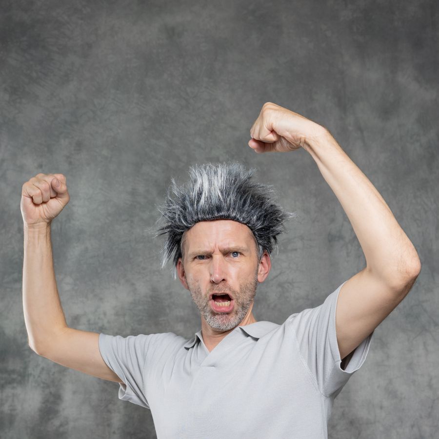 Een man met een pruik van grijs haar dat rechtop staat, staat voor een grijze achtergrond met zijn armen omhoog alsof hij spierballen laat zien en kijkt stoer.