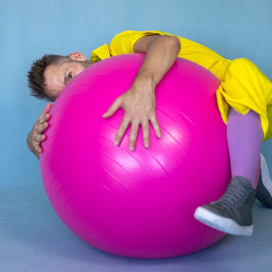Een man in een geel pak ligt half over een ronde roze bal met een blauwe achtergrond. 