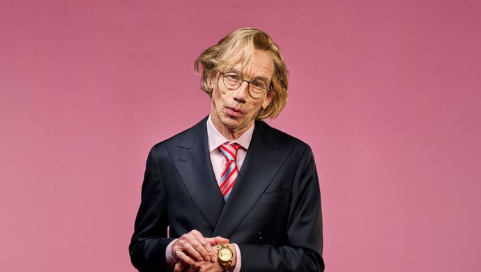 Een man in een net pak kijkt niet blij naar de camera terwijl hij zijn hand vasthoudt waar een horloge zit. Bij zijn benen zit een vrouw. Alleen haar handen om zijn heupen en haar haar in een knot is in beeld. De achtergrond is roze.