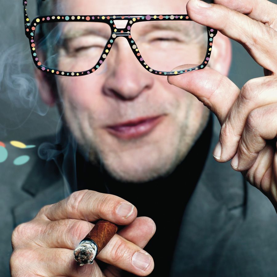 Erik houdt een bril en een sigaar vast. Hij heeft één oog dicht terwijl hij door de bril tuurt naar de camera.