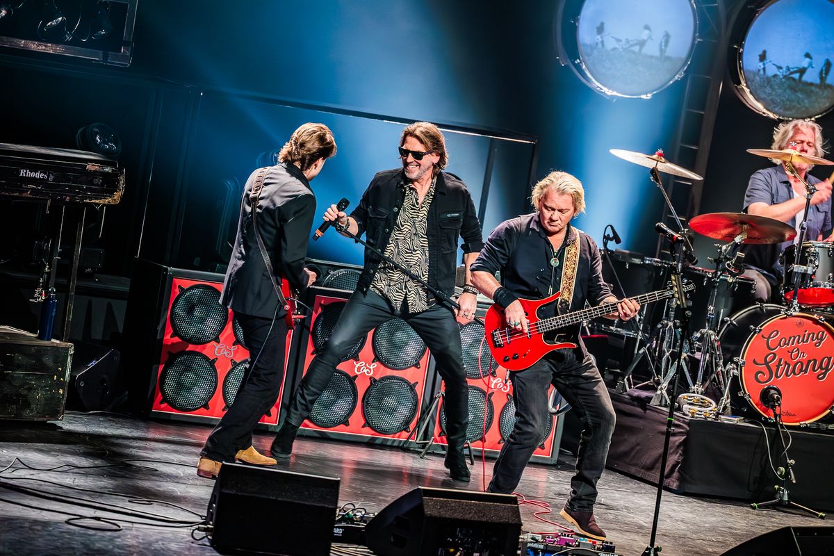 De bandleden rockend op het podium, de gitarist heeft een rode gitaar in zijn handen, de zanger houdt het statief van de microfoon in beide handen. 