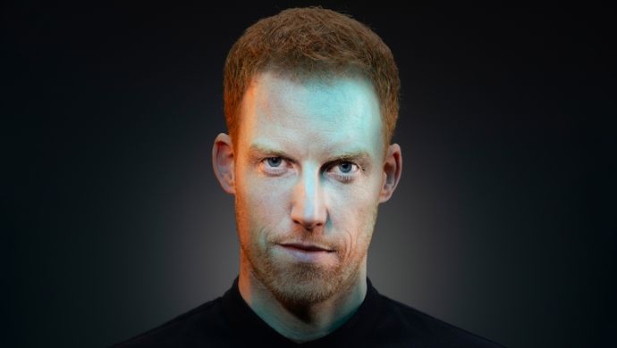 René kijkt serieus naar de camera. Er zit een blauwe gloed over zijn gezicht heen. De achtergrond is zwart en hij heeft een zwart shirt aan.