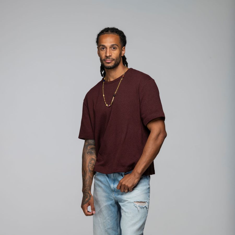 Jay staat voor een witte achtergrond in een spijkerbroek en een donkerrood shirt. hij heeft een hand los langs zijn lijf hangen en de andere hand bij zijn broekzak. Hij kijkt neutraal de camera in.