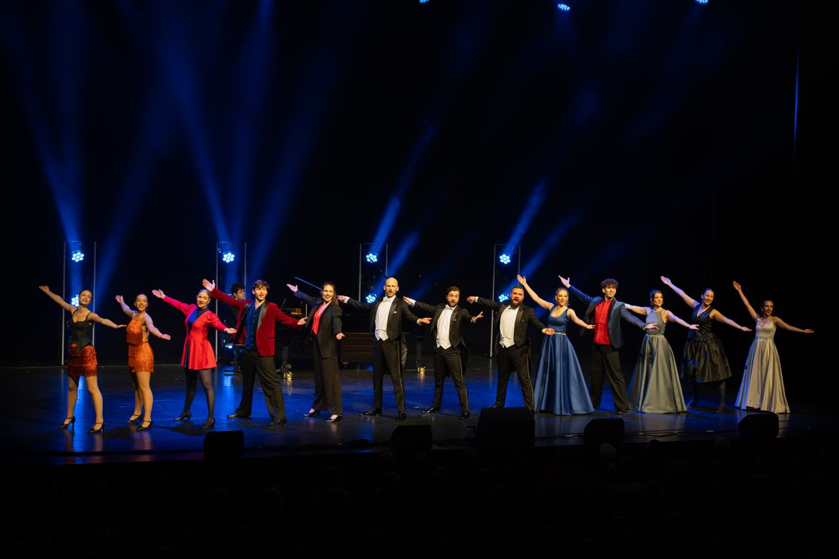 Een groep mensen staan op een podium en hebben allemaal hun armen wijd op dezelfde manier. Achter hun staan lampen die een blauwe kleur schijnen.
