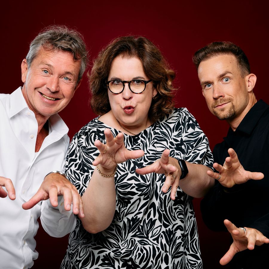 Drie mensen staan naast elkaar voor een donkerrode achtergrond. Ze kijken met een geheimzinnige blik recht vooruit en houden hun handen voor zich omhoog, met gespreide vingers.