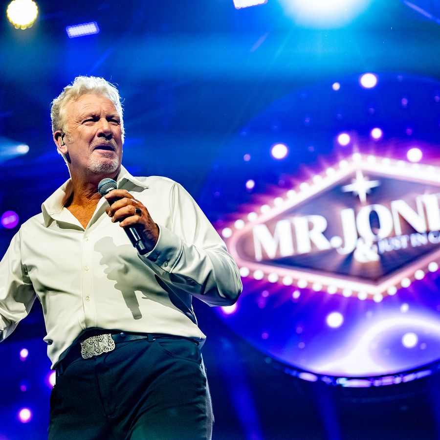 Een zanger kijkt het publiek in. Hij houdt een microfoon vast en zijn andere hand heeft hij een beetje omhoog. Hij staat op het podium met op de achtergrond de woorden: 'Mr. Jones & Just in Case'.