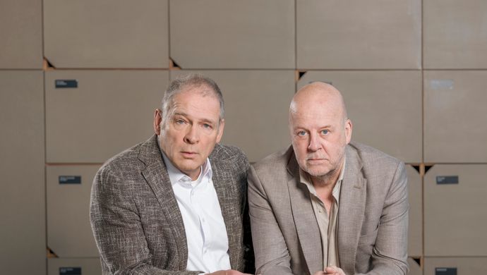 Twee mannen, kalend in grijze/beige jasjes leunen op een tafel, achter hen een rij lockerkastje. 