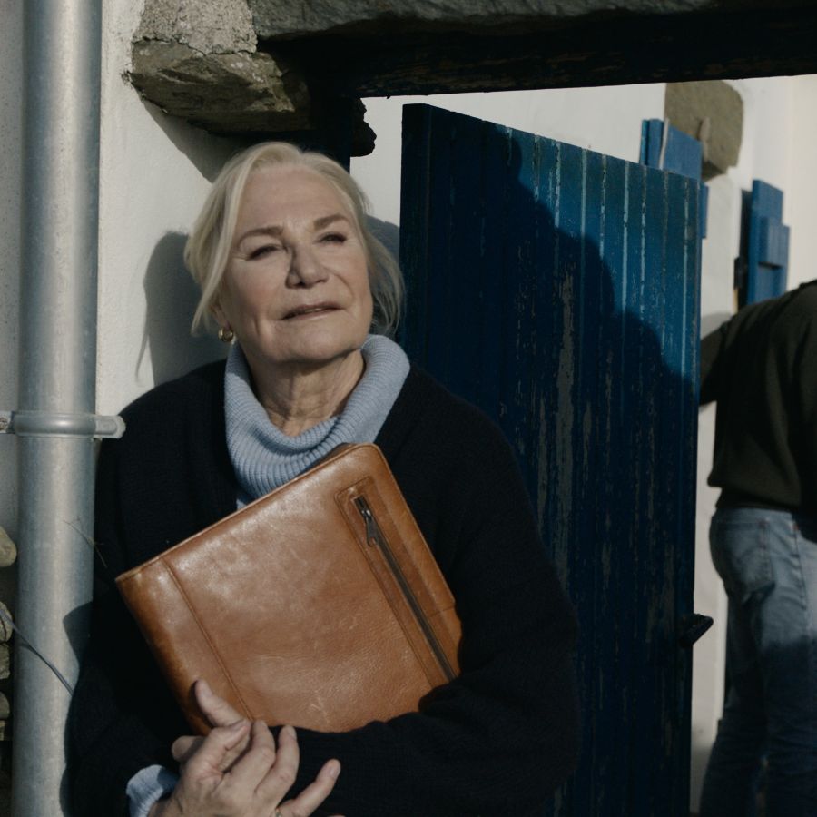 Een vrouw met tas in de armen staat in het zonlicht voor haar huis, met op de achtergrond een jonge man die de deur dicht doet en naar haar kijkt. 