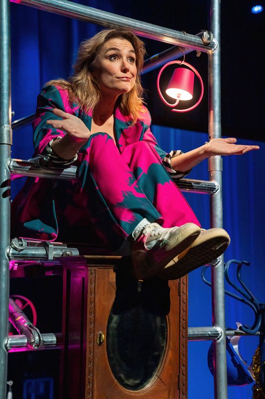 Evi Hansen zit met haar benen over stangen van een stellage. Ze kijkt vertwijfelend naar voren en haalt haar schouders op met haar armen wijd. Naast haar hangt een roze lampje. Ze heeft een roze met groene pak aan en de achtergrond is blauw.