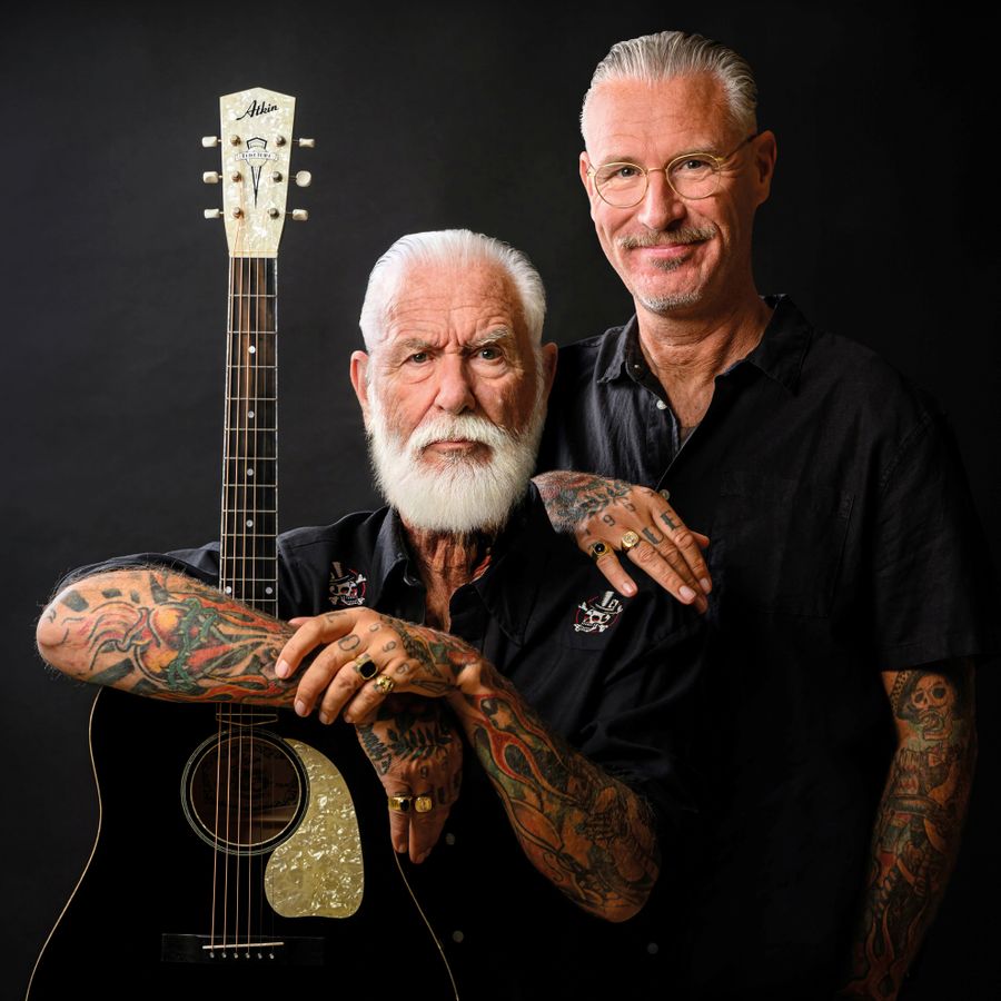 Twee mannen, die vader en zoon moeten voorstellen kijken recht in de camera. De vader heeft wit haar en een witte baard en omarmd zijn gitaar. Zijn zoon staat naast hem. Allebei hebben ze hun armen vol tatoeages. 