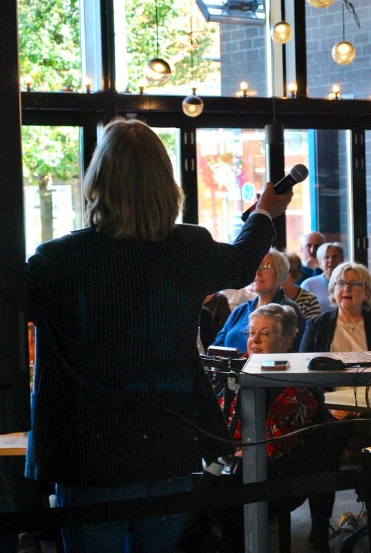 Een man staat met zijn rug naar de camera en houdt een microfoon vast. Op de achtergrond zie je een groep mensen zingen.