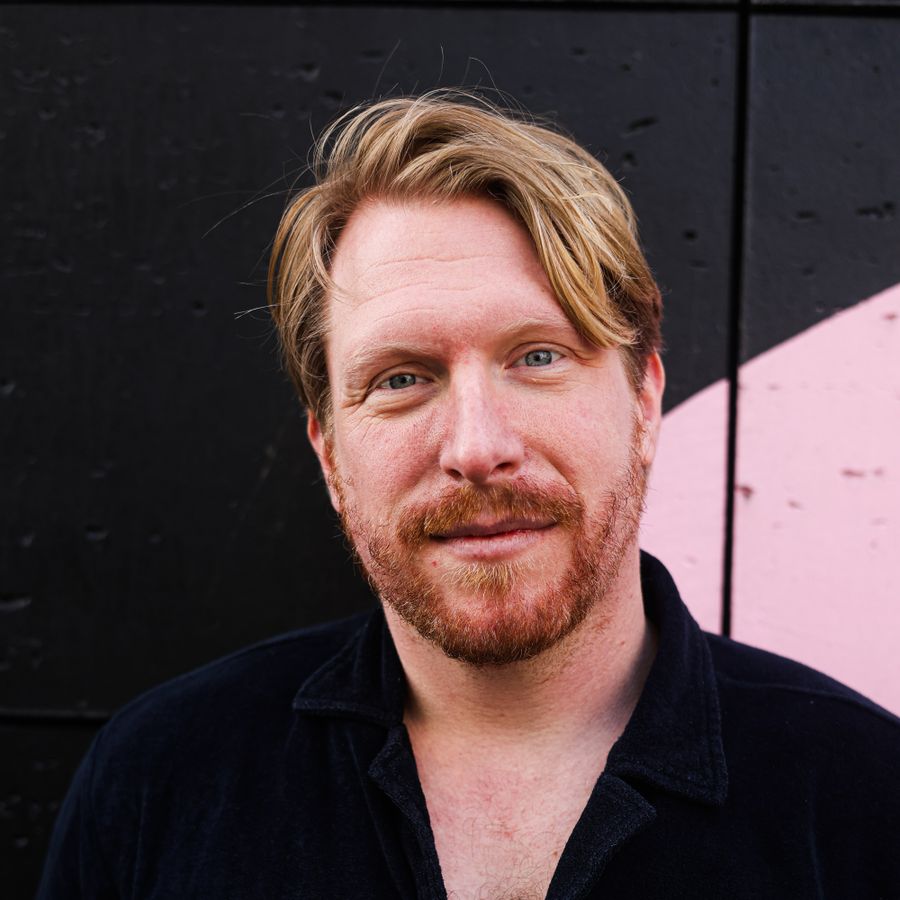Kor Hoebe staat voor een zwarte achtergrond met een beetje roze. Hij heeft een zwart shirt aan en houdt zijn handen op zijn rug. Hij kijkt glimlachend de camera in.