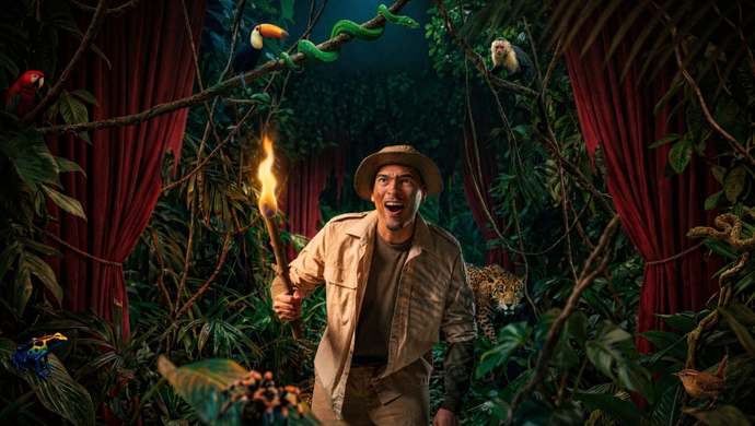 Man in survival outfit met een fakkel in zijn hand loopt door een jungle achtig decor met theatergordijnen. 