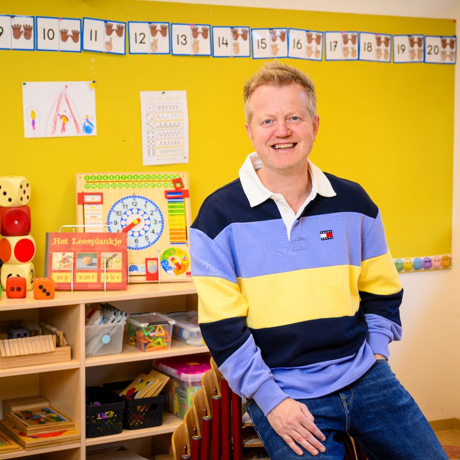 Meester mark zit in een klaslokaal. Hij leunt tegen een stapel stoelen met zijn hand in zijn zak. Op de achtergrond is een kast met speelgoed te zien.