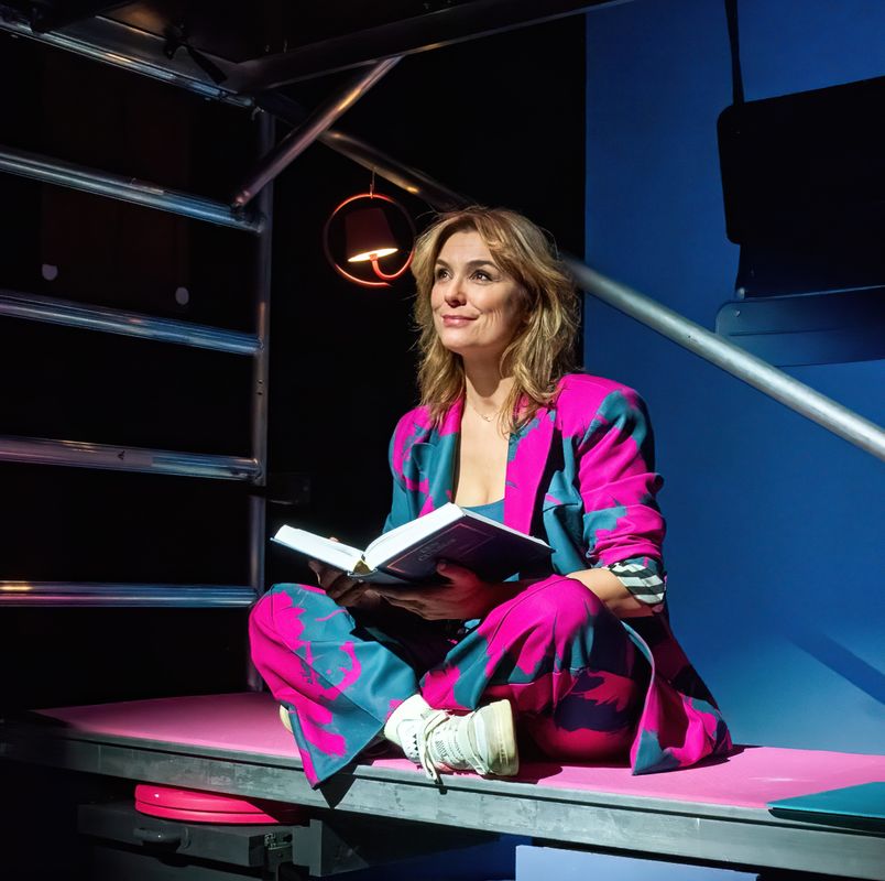 Evi Hansen zit op een stellage met een boek in haar handen. Ze kijkt met een glimlach naar voren en houdt het boek open. Ze heeft een roze met groene pak aan en de achtergrond is blauw.