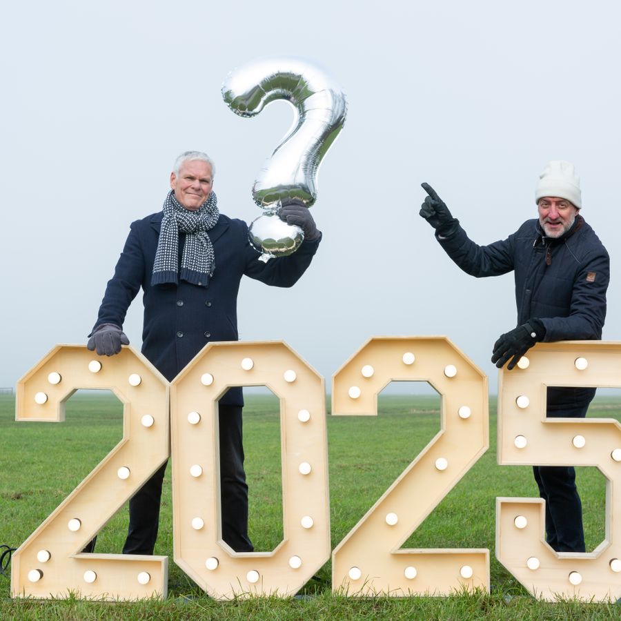 Rob en joep staan met winterkleding op een grasveld achter grote 2025 cijfers. De één heeft een vraagteken in zijn hand en de ander wijst naar dat vraagteken. Allebei lachen ze naar de camera.