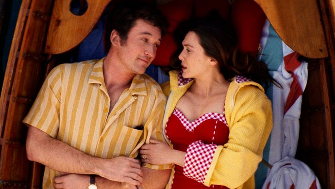 Man in geel overhemd ligt naast een vrouw in rood badpak met geel blouse, ze  kijken elkaar aan. 