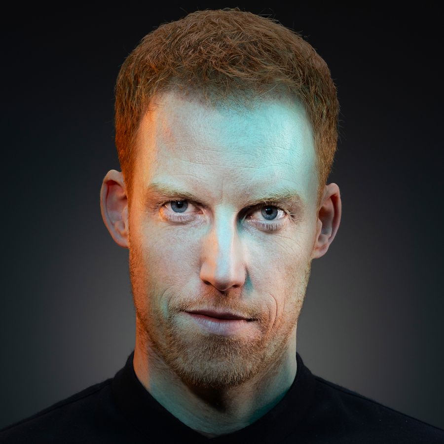 René kijkt serieus naar de camera. Er zit een blauwe gloed over zijn gezicht heen. De achtergrond is zwart en hij heeft een zwart shirt aan.