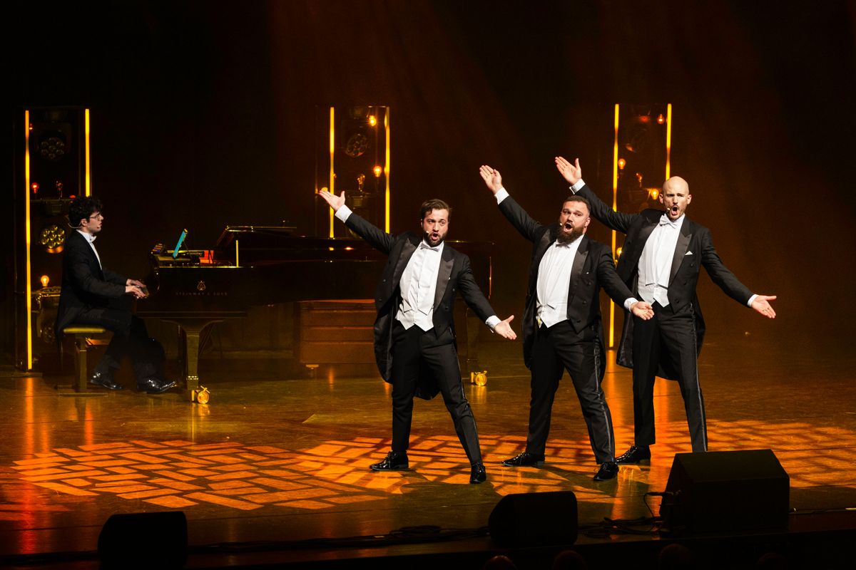 Drie mannen in pak staan op het podium met hun armen open alsof ze iets laten zien. Ze zingen en op de achtergrond is een pianist te zien.