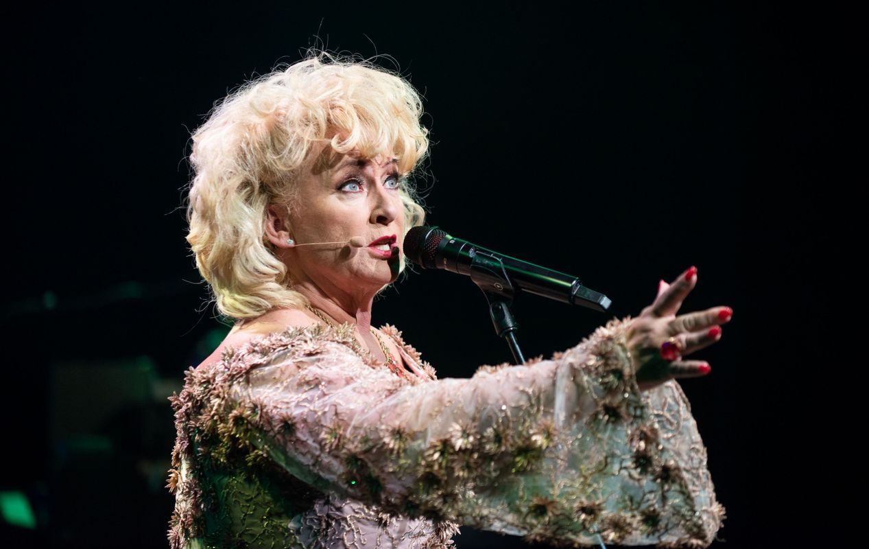 Karin Bloemen staat voor een microfoon en zingt. Ze heeft een roze jurk aan en houdt haar hand naar voren. Ze heeft een verdrietige of gevoelige uitdrukking op haar gezicht.