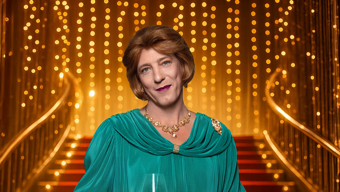 Marja staat in een blauwgroene jurk met een glas wijn in haar hand. Ze kijkt glimlachend naar de camera en staat voor een trap met een rode loper en een goude reling. Boven aan de trap zijn goude lichtjes te zien.