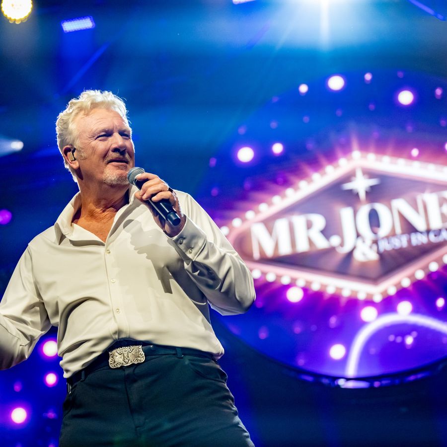 Mr Jones staat met een microfoon op een podium. Achter hem hangt een grote lamp met Mr. Jones erop.