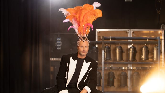 Man in chique smokingpak met witte accenten zit op toneel met op achtergrond theaterlampen in een rek. Op zijn hoofd draagt hij een verentooi welke showgirls dragen. 