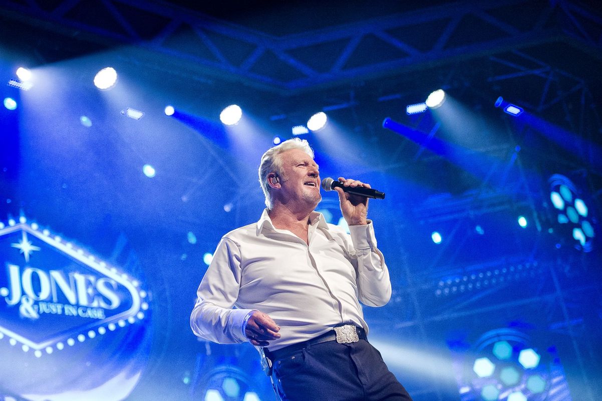 Mr Jones staat met een microfoon in zijn hand en zingt erdoorheen. Op de achtergrond zijn blauwe lampen te zien.