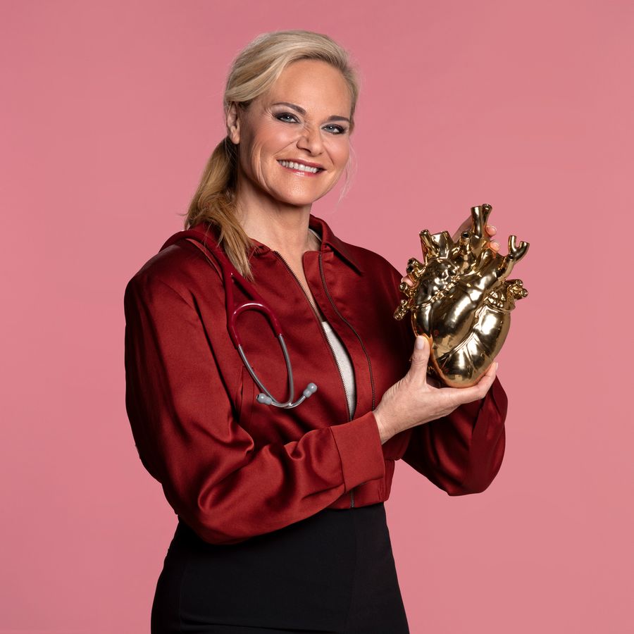 Vrouw met roodroze blouse en zwarte broek houdt een gouden mensenhart vast. Ze kijkt lachend in de camera. 
