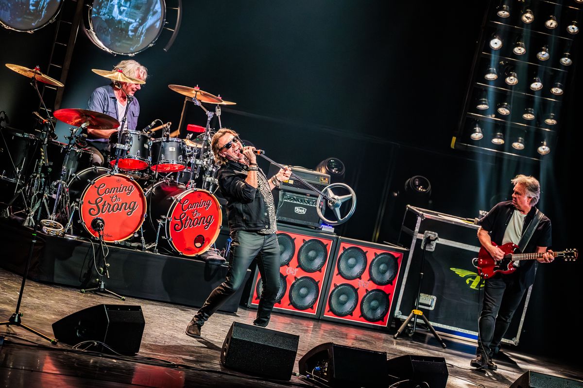 Zanger zingt uit volle borst met op achtergrond de drummer en rechts van hem  de gitarist. De zanger buigt ver naar achter en tilt de microfoon omhoog.