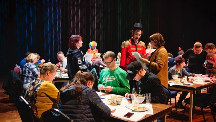 Er zitten allemaal groepjes mensen aan tafel met allemaal papieren op tafel. Midden in de groep loopt een man rond in een rood pak met een zwarte hoge hoed.