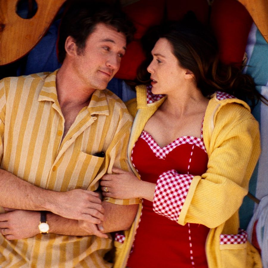 Man in geel overhemd ligt naast een vrouw in rood badpak met geel blouse, ze  kijken elkaar aan. 