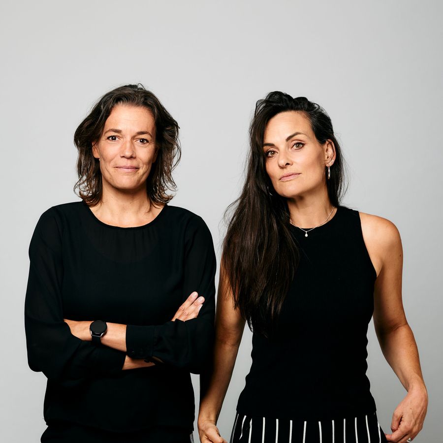 Twee vrouwen zijn zwart gekleed en staan voor een grijze achtergrond. De vrouw links staat met haar armen over elkaar en de vrouw rechts heeft haar armen langs haar lichaam. Ze kijken allebei met een glimlach de camera in.