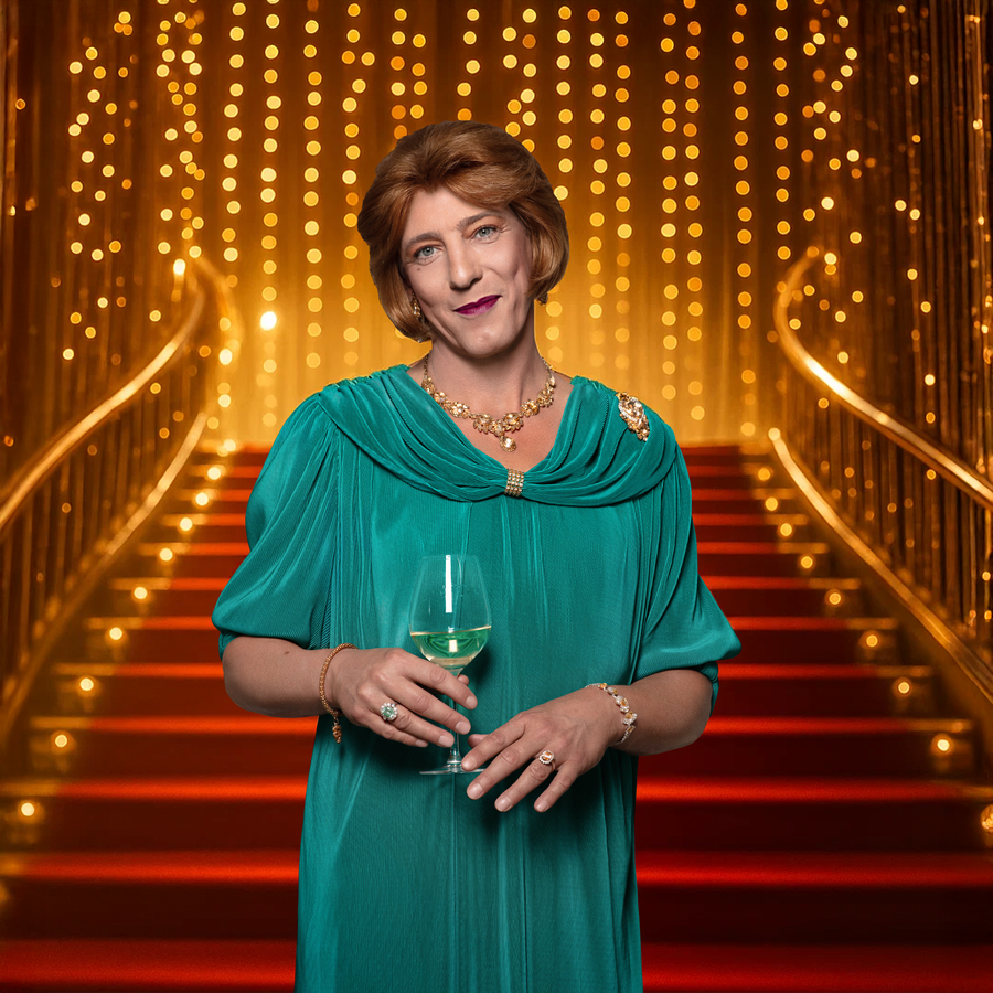 Marja staat in een blauwgroene jurk met een glas wijn in haar hand. Ze kijkt glimlachend naar de camera en staat voor een trap met een rode loper en een goude reling. Boven aan de trap zijn goude lichtjes te zien.