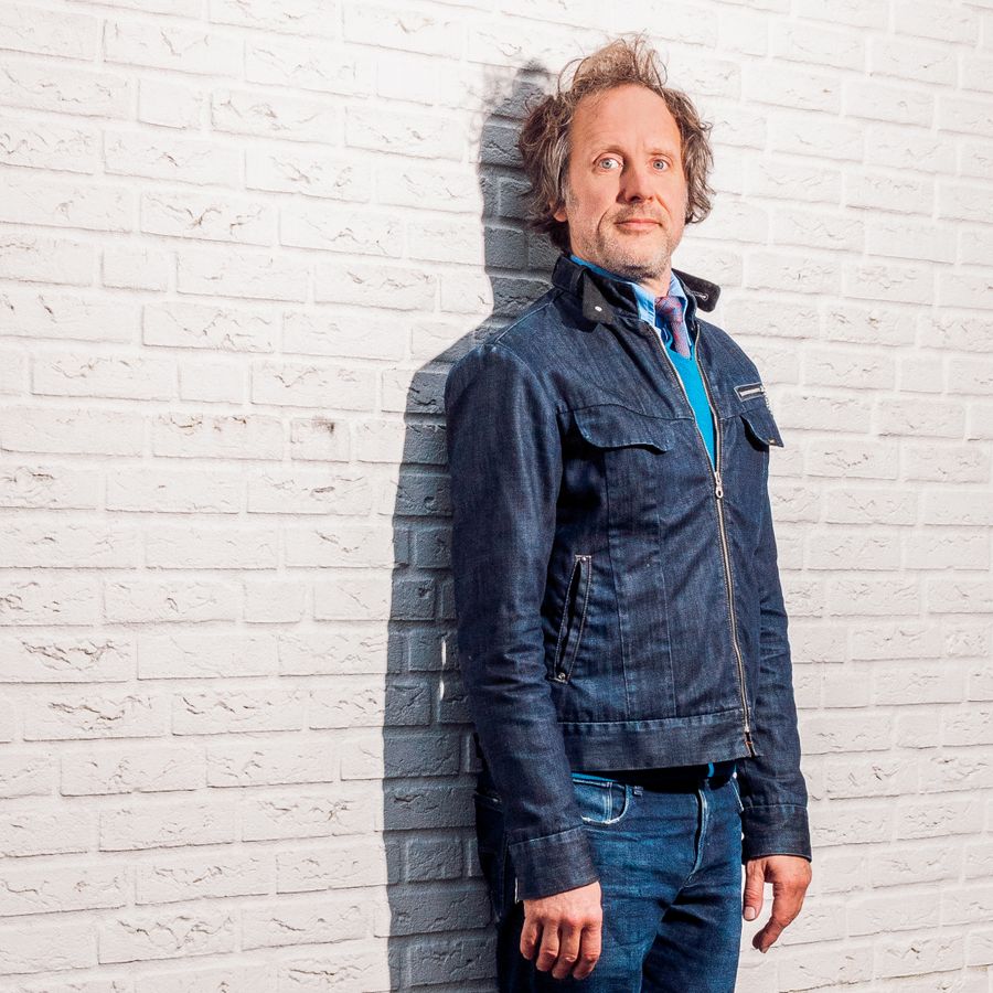 Ronald staat tegen een witte muur. Hij kijkt serieus naar de camera en staat met zijn armen langs zijn lijf, stijf tegen de muur aan.