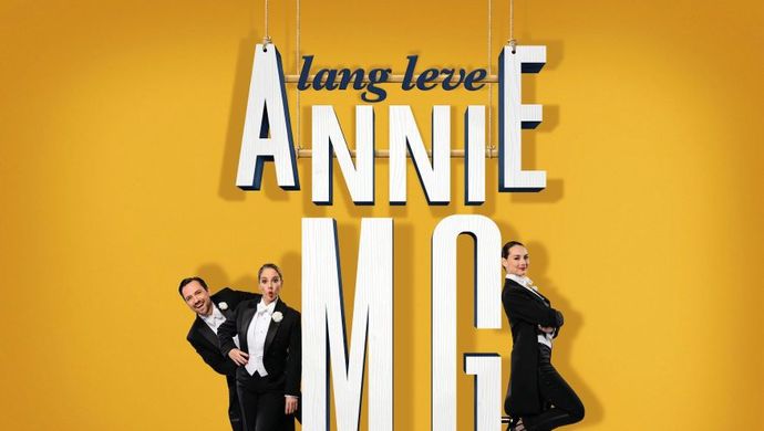 Een geel plaatje met witte letters: lang leve Annie M.G. met drie mensen in een zwart pak. Twee vrouwen en een man.