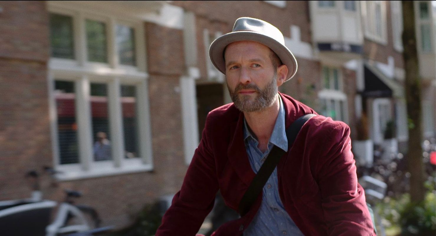 Man met rood jasje en tas over de schouder fietst door een straat. 