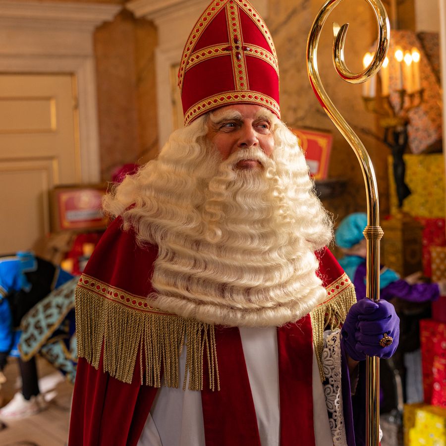 Sinterklaas met staf in de hand kijkt naar iemand in de deuropening. 