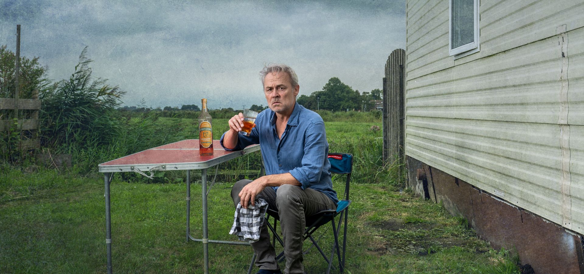 Marcel zit naast een muurtje in een soort weiland aan een campingtafeltje op een campingstoel. Hij houdt een theedoek in zijn handen en drinkt uit een glas. Op tafel naast hem staat een fles drank. Hij kijkt ernstig de camera in.