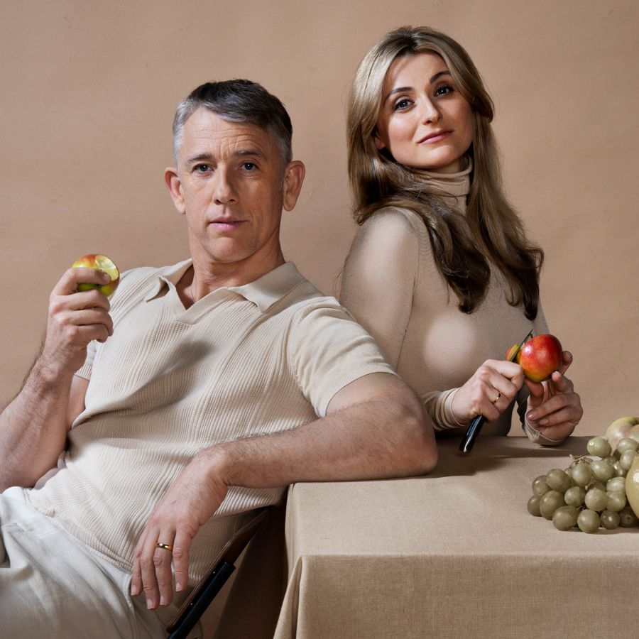 Een man en een vrouw zitten leunend aan een tafel. De man eet een appel en kijkt serieus naar de camera. De vrouw snijdt een appel en kijkt ook serieus naar de camera. Op tafel ligt er groen fruit en de tafel, de achtergrond en hun kleding is beige.