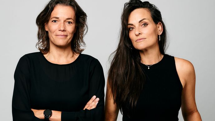 Twee vrouwen zijn zwart gekleed en staan voor een grijze achtergrond. De vrouw links staat met haar armen over elkaar en de vrouw rechts heeft haar armen langs haar lichaam. Ze kijken allebei met een glimlach de camera in.