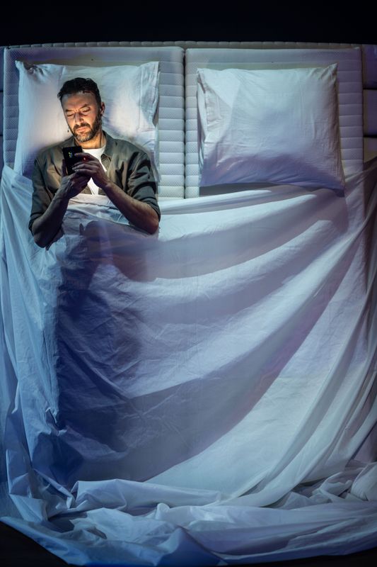 Man liggend in een witbed onder een wit laken met zijn armen gekruist op zijn lichaam. 