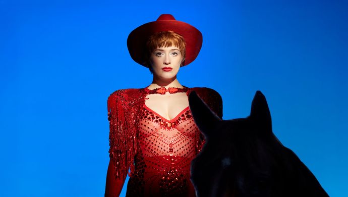 Er staat een vrouw en een paard voor een blauwe achtergrond. De vrouw heeft een rode top aan met gaas zodat je haar buik ziet en ze heeft een rode hoed op.